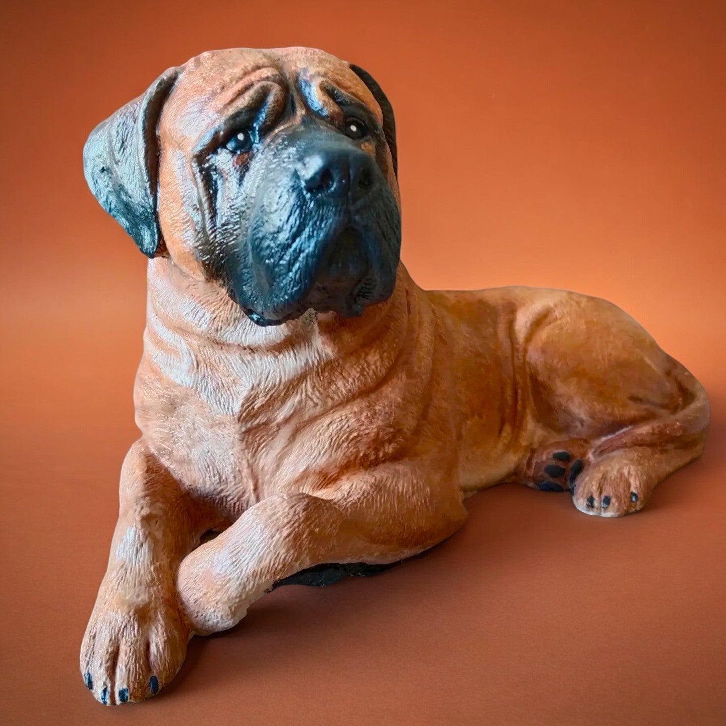 Bull Mastiff statue made of concrete and hand painted. Dog statue is laying down. Carmel brown colour with black snout and ears. Front view of statue