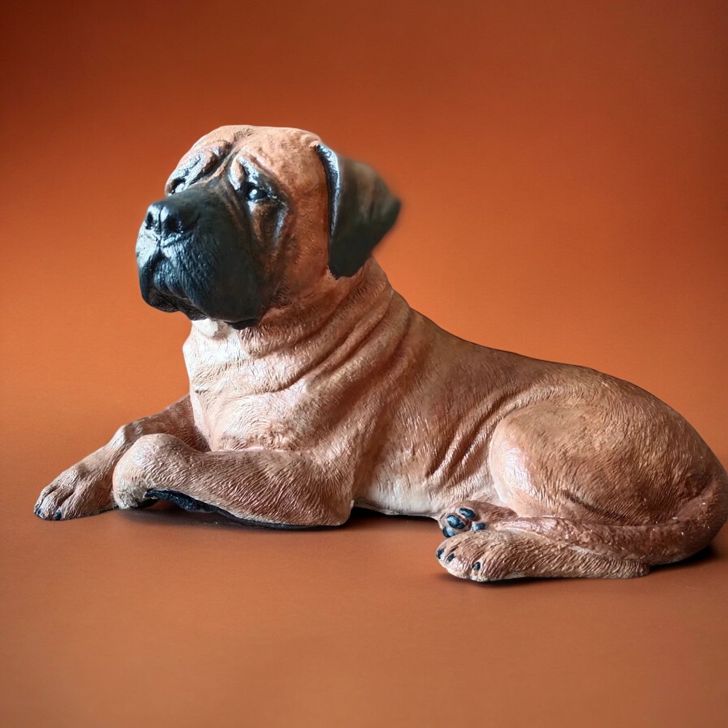 Bull Mastiff statue made of concrete and hand painted. Dog statue is laying down. Carmel brown colour with black snout and ears. Left side of statue